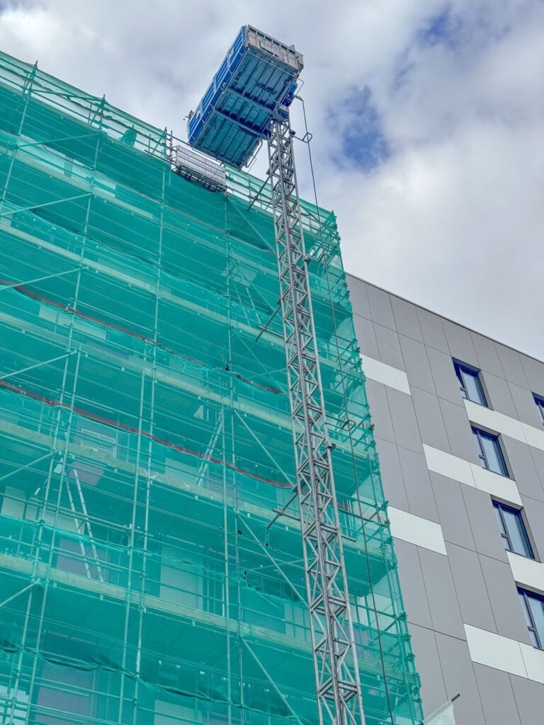 Échafaudage de façade couvert de filet vert, avec un lift de chantier MABER arrivé en haut de son mât